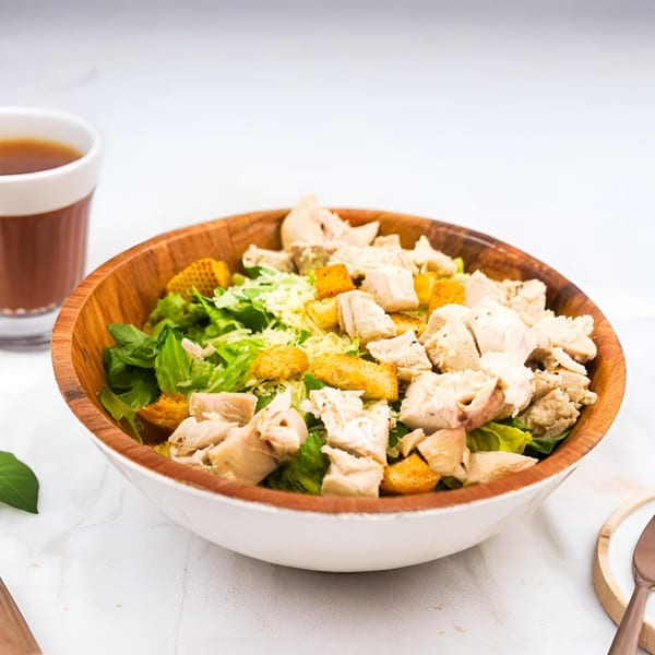 A wooden bowl of Ribline BBQ chicken Caesar salad with croutons and a drink.