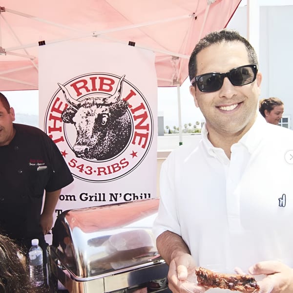 A man in sunglasses smiles, holding a Ribline BBQ rib, with a chef and banner behind him.