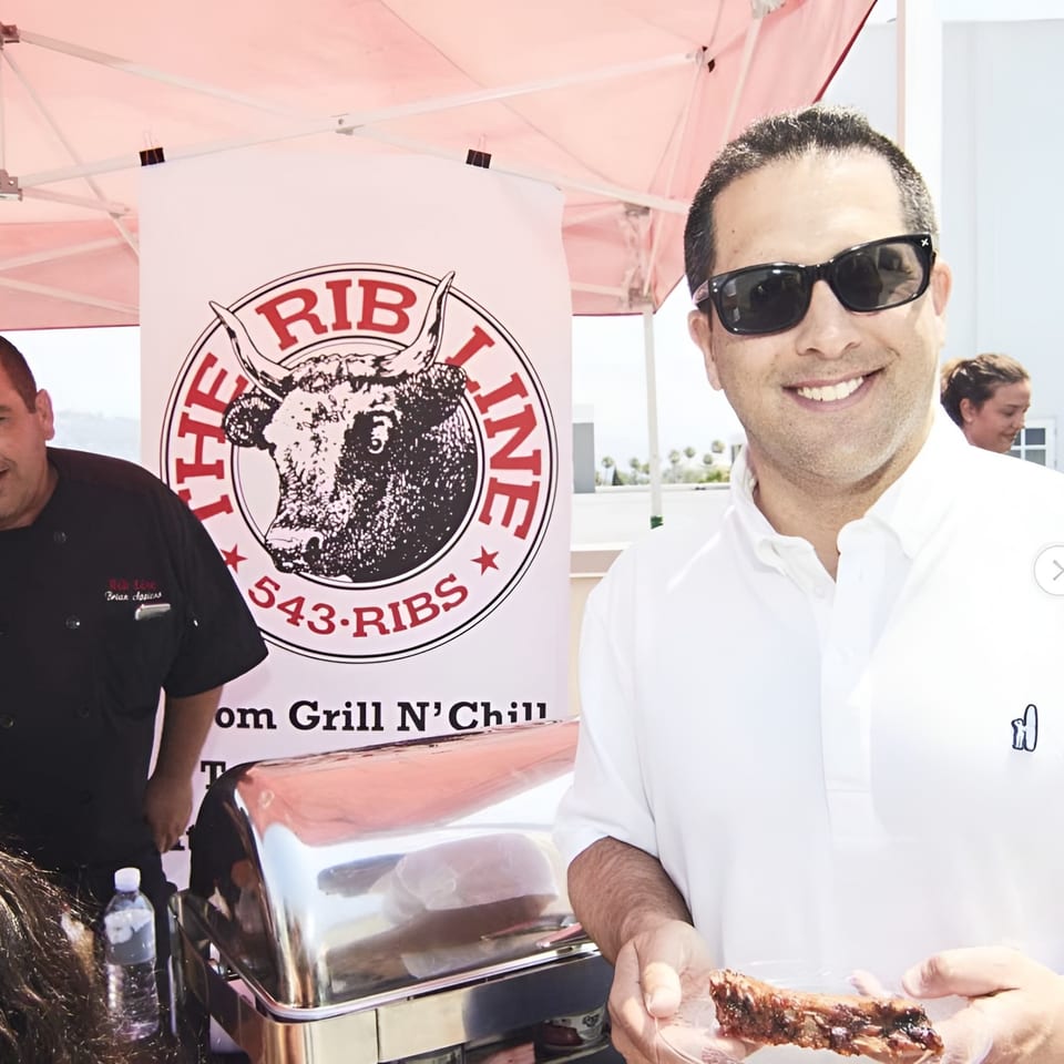 A man in sunglasses smiles, holding a Ribline BBQ rib, with a chef and banner behind him.