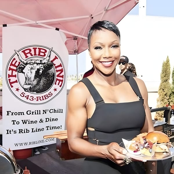 Smiling woman in black dress holds a plate of Ribline BBQ food next to a branded banner.