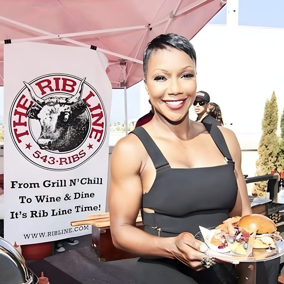 Smiling woman in black dress holds a plate of Ribline BBQ food next to a branded banner.