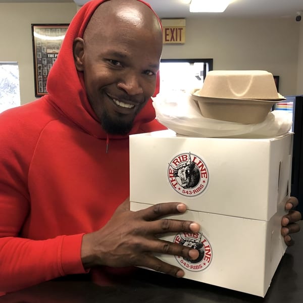 Jamie Foxx smiling, holding two Ribline BBQ boxes and a takeout container.